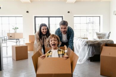 Happy parents play with their excited son in a cardboard box while packing their home for moving abroad for work with a young child.