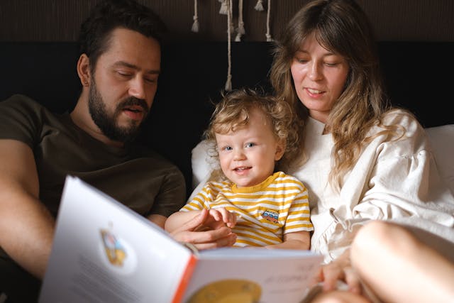 two adults and a child sitting closely together, reading a book with colorful illustrations